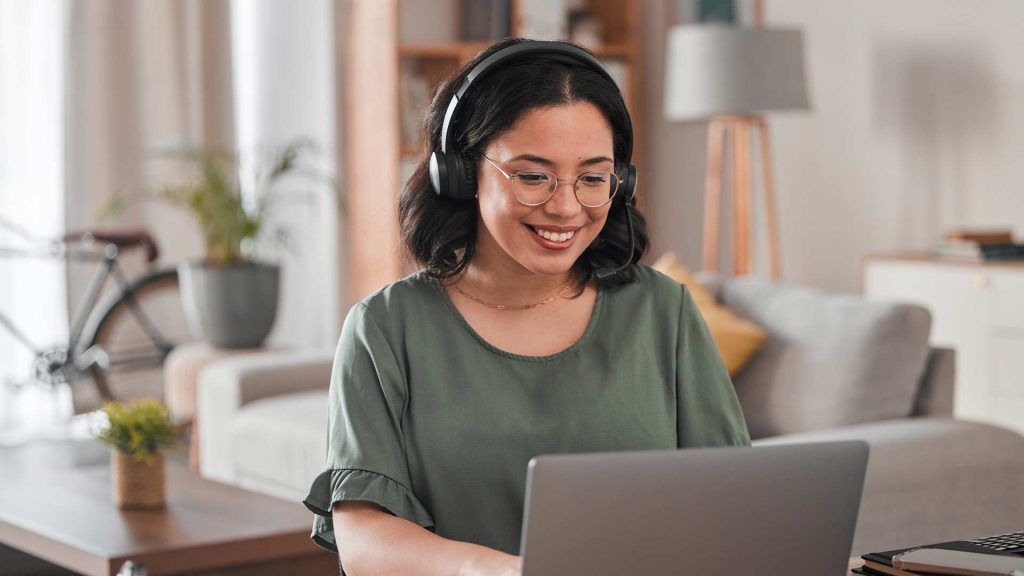 The image depicts a young woman, who is sitting at a desk using a laptop computer. She appears to be working from home or in a comfortable, cozy office setting. This image would be an appropriate representation for the keyword "best place to hire virtual assistant", as it depicts the type of skilled, professional individual who could fulfill that role.